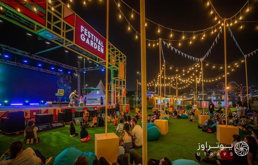 مردم در محوطه‌ای چمن‌کاری‌شده مقابل سن اجرای برنامه در فستیوال غذای دبی، نشسته‌اند. 